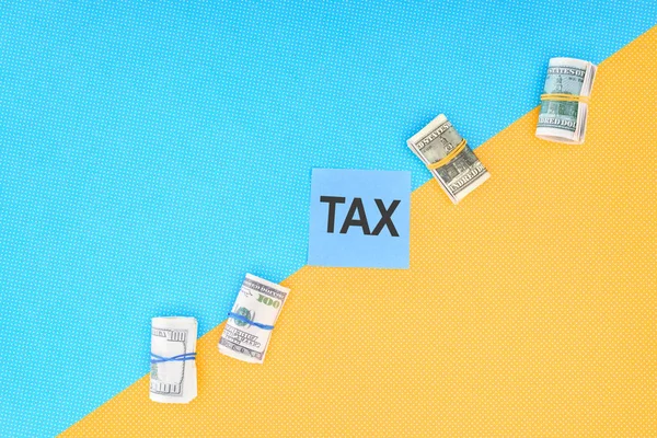 Vista superior de rollos de dinero y tarjeta con la palabra 'impuesto' sobre fondo azul y amarillo - foto de stock