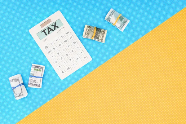 Top view of calculator with word 'tax' and money rolls on blue and yellow background — Stock Photo