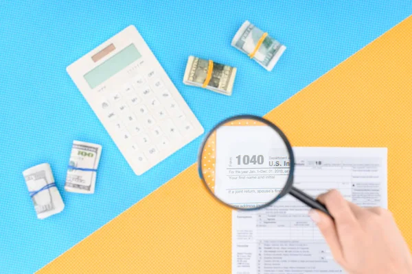 Vista recortada de la mujer sosteniendo lupa sobre el formulario de impuestos sobre fondo azul y amarillo - foto de stock