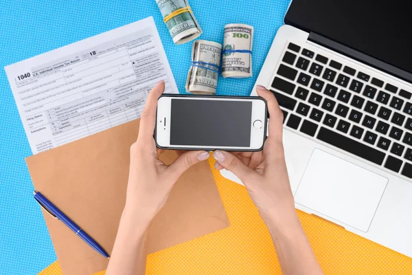 Vista parcial de la mujer sosteniendo teléfono inteligente con pantalla en blanco en el lugar de trabajo con computadora portátil, formulario de impuestos y rollos de dinero en segundo plano - foto de stock