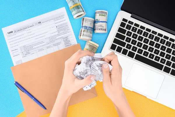Cropped view of woman holding crumpled paper ball at desk with tax form, laptop and money rolls — Stock Photo
