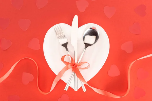 Верхний вид тарелки со столовыми приборами, обернутыми праздничной лентой рядом с символами сердца, изолированными на красный день святого Валентина — стоковое фото