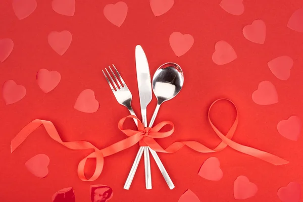 Vista elevada de los cubiertos envueltos por la cinta festiva cerca de símbolos del corazón aislados en rojo, San Valentín concepto de día - foto de stock