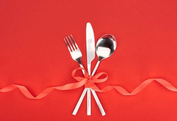 Vista superior de tenedor, cuchillo y cuchara envueltos por cinta festiva roja aislada en rojo, concepto de San Valentín - foto de stock
