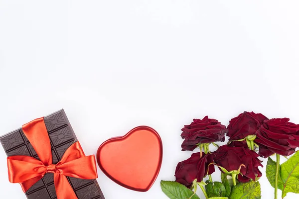 Повышенный вид шоколада, завернутый праздничной лентой, красные розы и сердце форме подарочной коробки изолированы на белом, День святого Валентина концепции — стоковое фото