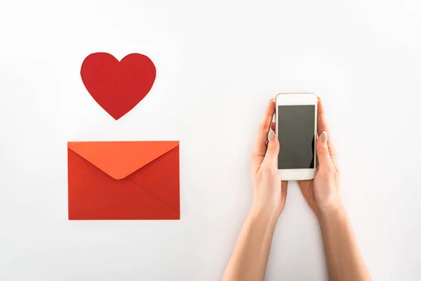 Teilansicht der Frau mit Smartphone mit leerem Bildschirm in der Nähe des Herzsymbols und rotem Umschlag auf weißem, Valentinstag-Konzept — Stockfoto