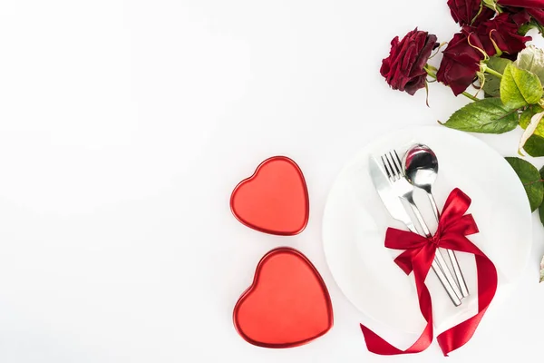 Ansicht von Teller und Besteck umhüllt von festlicher Schleife, herzförmigen Geschenkschachteln und Strauß roter Rosen isoliert auf weißem, Valentinstag-Konzept — Stockfoto