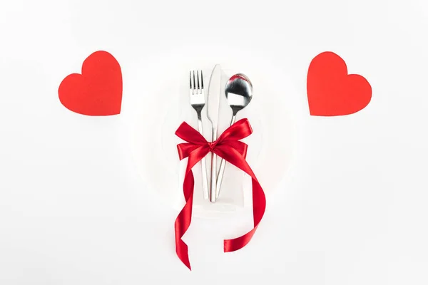 Elevated view of plate with cutlery wrapped by festive ribbon bow near heart symbols isolated on white, st valentine day concept — Stock Photo