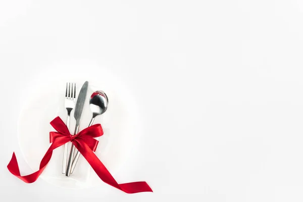 Vista elevada de tenedor, cuchillo y cuchara envueltos por arco festivo rojo en la placa aislada en blanco, San Valentín concepto de día - foto de stock