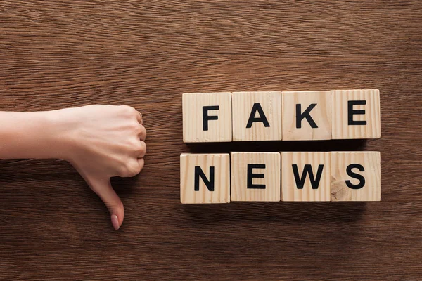 Image recadrée de journaliste montrant pouce vers le bas à cubes en bois avec des mots fausses nouvelles à la table en bois — Photo de stock