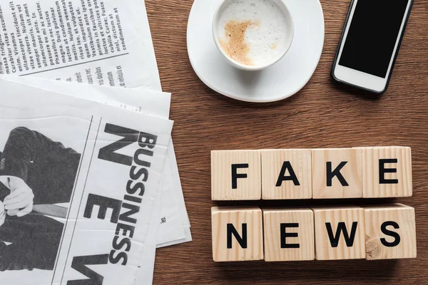 Top view of wooden cubes with words fake news, cup of coffee and smartphone on wooden tabletop — Stock Photo