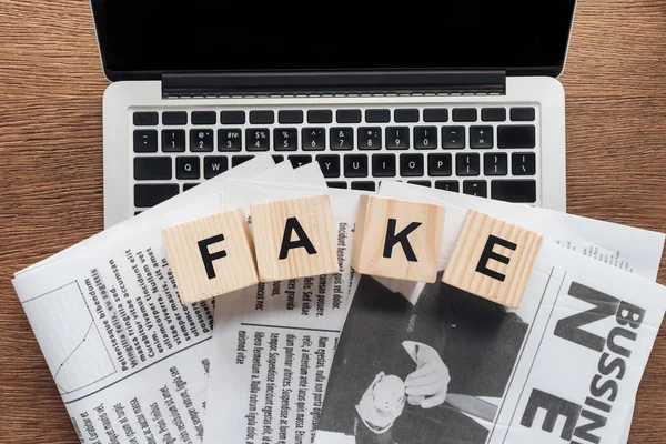 Top view of wooden cubes with word fake and newspapers on laptop on wooden tabletop — Stock Photo