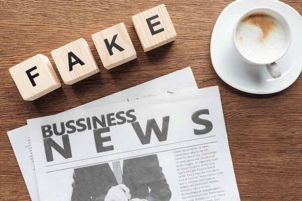 Top view of wooden cubes with word fake, business newspaper and cup of coffee on wooden tabletop — Stock Photo