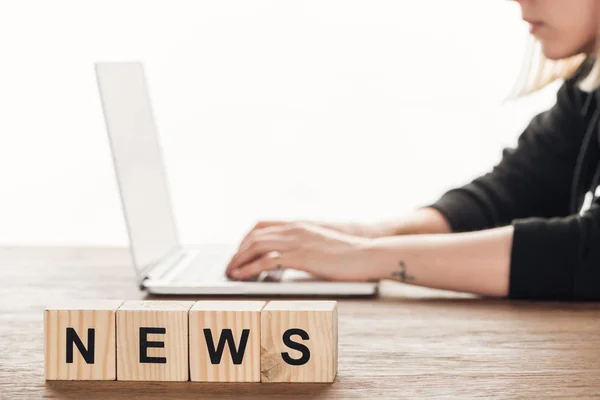Обрезанное изображение журналиста, работающего за ноутбуком, деревянные кубики с новостями за деревянным столом — стоковое фото