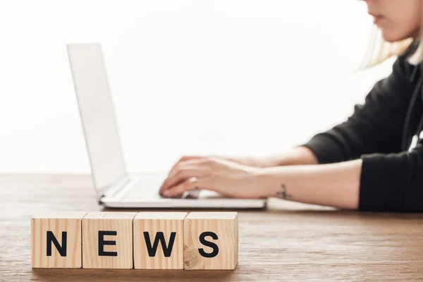 Imagen recortada de periodista que trabaja en el ordenador portátil, cubos de madera con noticias de palabra en la mesa de madera - foto de stock