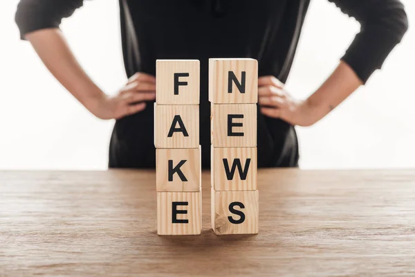 Cropped image of journalist standing with hands akimbo, wooden cubes with words fake news at wooden table — Stock Photo