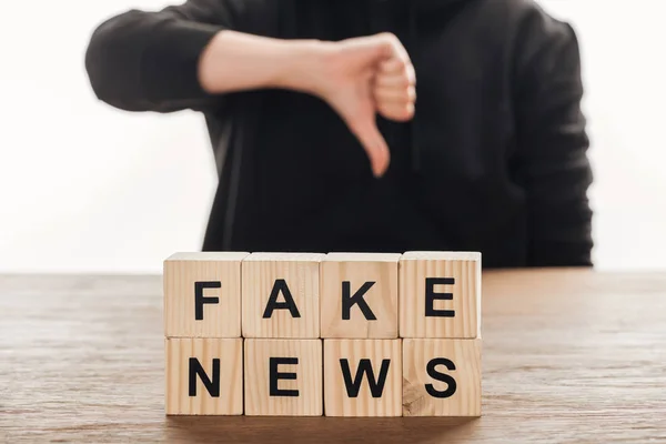 Image recadrée de journaliste montrant pouce vers le bas à cubes en bois avec des mots fausses nouvelles à la table en bois — Photo de stock