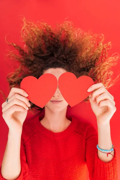 Vista superior de la niña sosteniendo tarjetas en forma de corazón en frente de la cara aislada en rojo, San Valentín concepto de día - foto de stock
