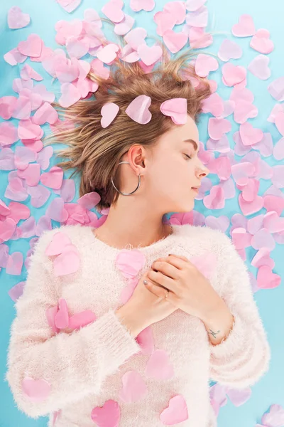 Top view of beautiful girl with eyes closed lying with heart shaped confetti isolated on blue, st valentines day concept — Stock Photo