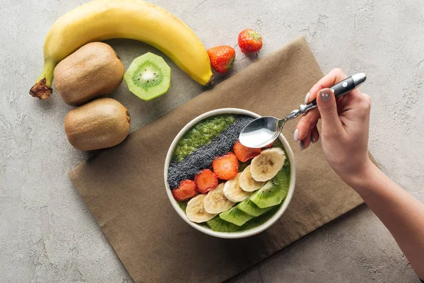 Vista superior de la mujer sosteniendo cuchara sobre un tazón de batido con frutas frescas sobre fondo gris - foto de stock