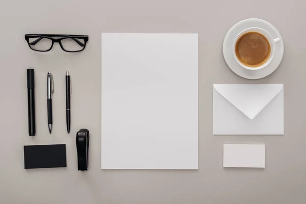 Tumbado plano con vasos negros, papelería y tarjetas blancas vacías en el lugar de trabajo con taza de café sobre fondo gris - foto de stock