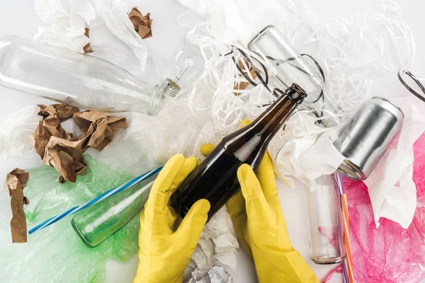 Vue recadrée de l'homme tenant une bouteille en verre vide parmi la boîte, les sacs en plastique, les bandes de papier, le papier et les tubes en plastique — Photo de stock