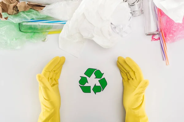 Vista parcial do homem em luvas de borracha amarela entre garrafas de vidro, sacos de plástico, papel e tubos de plástico com símbolo de reciclagem — Fotografia de Stock