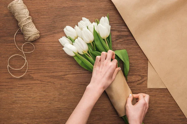 Vista ritagliata su fiorista facendo bouquet di tulipani bianchi, spago e carta artigianale su sfondo di legno — Foto stock