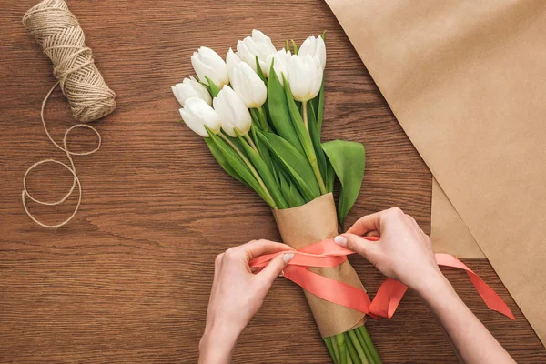 Vista recortada de floristería haciendo ramo de tulipanes blancos de primavera y cinta sobre fondo de madera - foto de stock