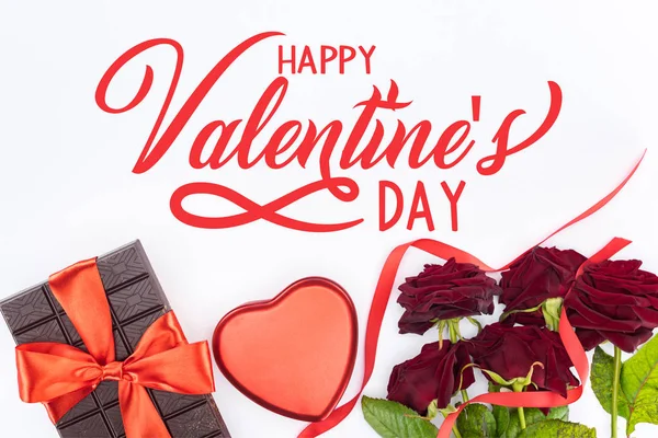Vue du dessus du chocolat enveloppé d'un ruban festif, de roses rouges et d'une boîte cadeau en forme de coeur isolée le jour de la Saint-Valentin blanche et heureuse — Photo de stock