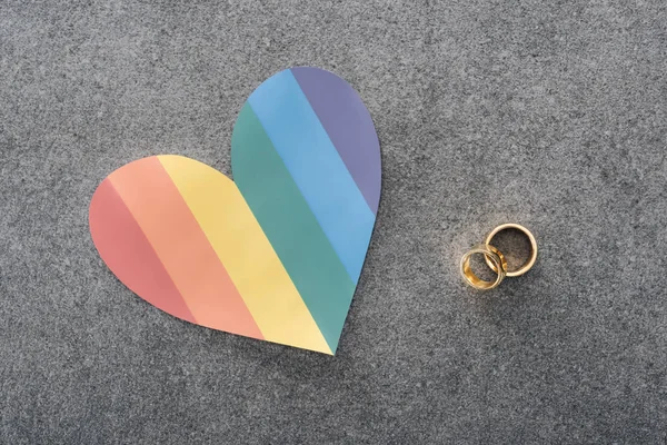 Arco-íris coração de papel colorido e anéis de casamento no fundo cinza, conceito lgbt — Fotografia de Stock