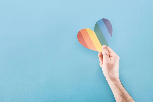 Vista cortada da mão masculina com coração de papel colorido arco-íris no fundo azul, conceito lgbt — Fotografia de Stock