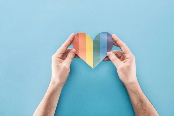 Vista recortada de manos masculinas con corazón de papel de color arco iris sobre fondo azul, concepto lgbt - foto de stock