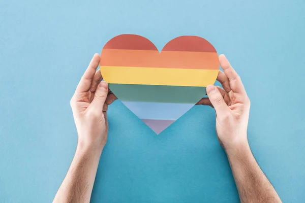 Partial view of man holding rainbow colored paper heart on blue background, lgbt concept — Stock Photo