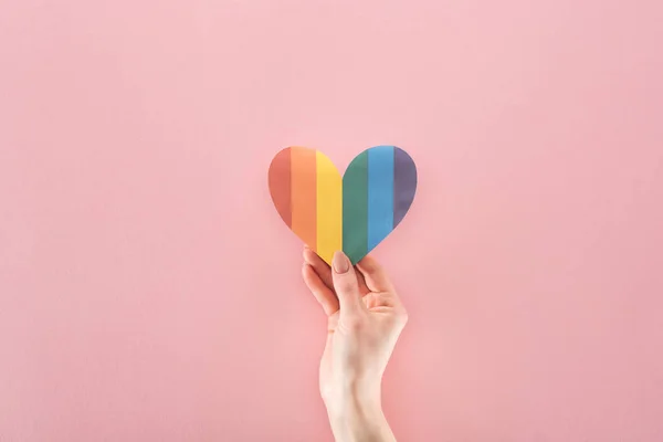 Vista recortada de la mano femenina con corazón de papel de color arco iris sobre fondo rosa, concepto lgbt - foto de stock