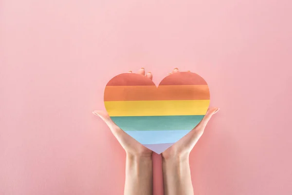 Vista parcial de la mujer sosteniendo el corazón de papel de color arco iris sobre fondo rosa, concepto lgbt - foto de stock