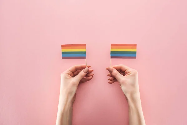 Partial view of female hands with paper rainbow flags on pink background, lgbt concept — Stock Photo