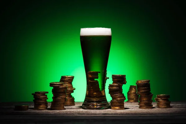 Vidro de cerveja em pé sobre mesa de madeira perto de moedas de ouro no fundo verde — Fotografia de Stock