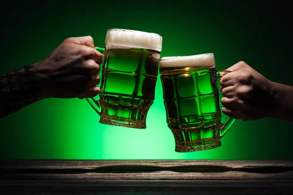 Vista recortada de amigos tintineando con vasos de cerveza irlandesa sobre fondo verde - foto de stock