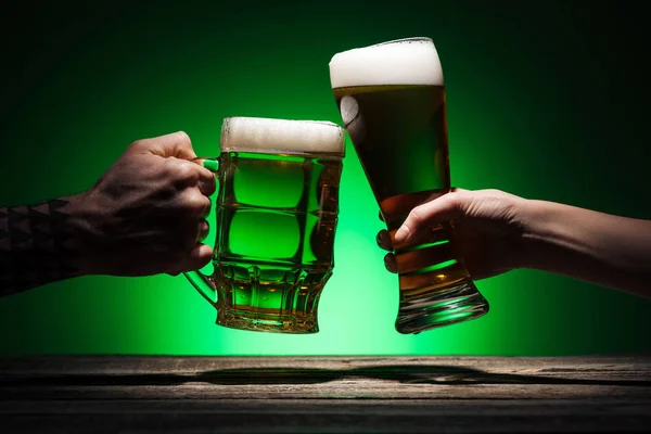 Vista recortada de los hombres tintineo con vasos de cerveza sobre fondo verde - foto de stock