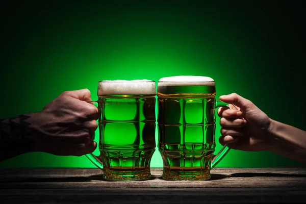 Vista recortada de amigos sosteniendo vasos de cerveza en st patricks día sobre fondo verde - foto de stock