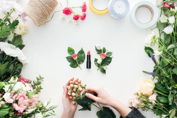 Vue recadrée des mains féminines tenant des baies sur les boutonnières et du rouge à lèvres entouré de fleurs sur fond blanc — Photo de stock