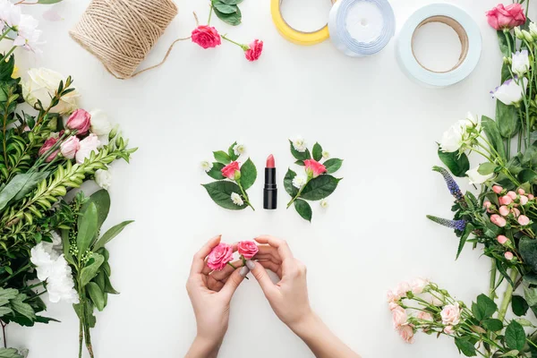 Vista ritagliata di mani femminili che tengono boccioli di rosa su boutonnieres e rossetto circondato da fiori su sfondo bianco — Foto stock