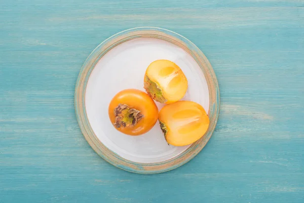 Top view of whole and sliced persimmons on glass plate and on blue background — Stock Photo