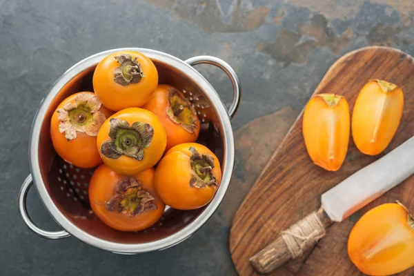 Vue de dessus de kaki orange en passoire et tranches sur planche à découper avec couteau — Photo de stock