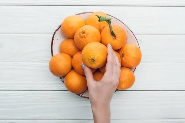 Vista ritagliata della mano femminile vicino piatto con mandarini su sfondo bianco in legno — Foto stock