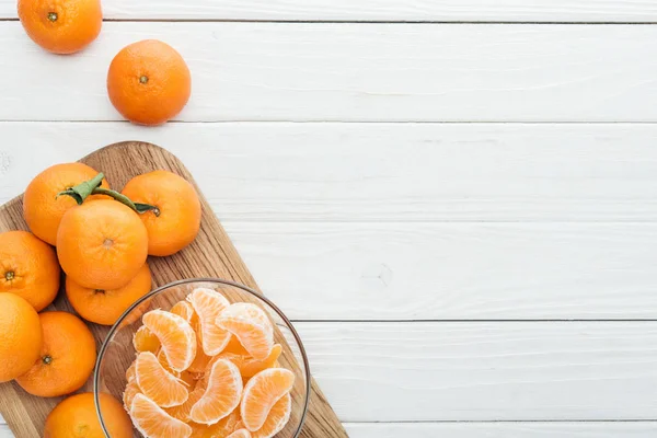 Vista superior de fatias de tangerina descascadas em tigela de vidro e tangerinas maduras inteiras em tábua de corte de madeira — Fotografia de Stock