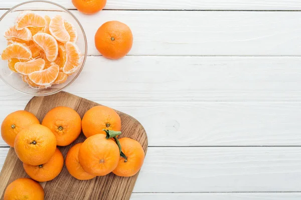 Vista superior de fatias de tangerina descascadas em tigela de vidro e tangerinas maduras inteiras em tábua de corte de madeira — Fotografia de Stock