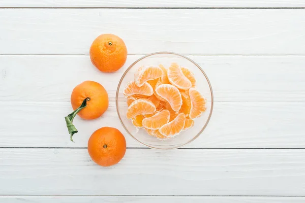 Vista superior de fatias de tangerina descascadas em tigela de vidro e tangerinas maduras inteiras em torno de fundo branco de madeira — Fotografia de Stock
