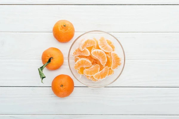 Top view of peeled tangerine slices in glass bowl and whole ripe tangerines around on wooden white background — Stock Photo